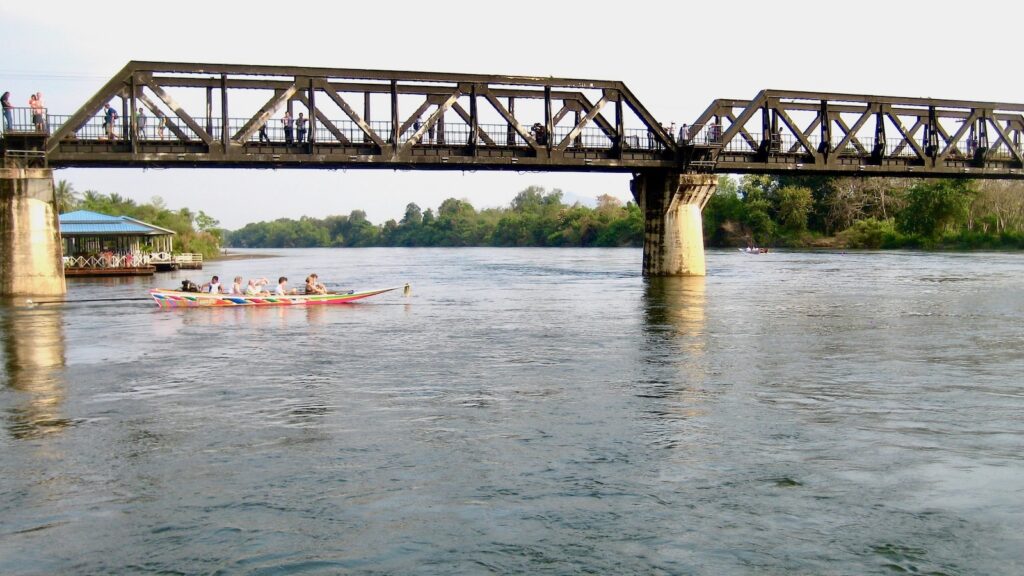 Visitors walking across the River Kwai Bridge in Kanchanaburi as a long-tail boat passes below